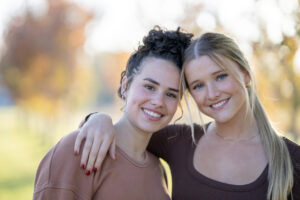 Two teen girls huddle in closely together outside, with their arms around one another, as they pose for a portrait. They are both dressed casually and are smiling.