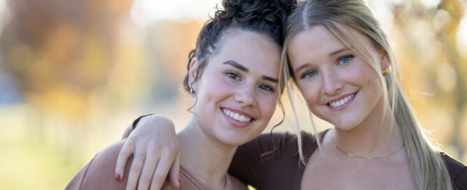 Two teen girls huddle in closely together outside, with their arms around one another, as they pose for a portrait. They are both dressed casually and are smiling.