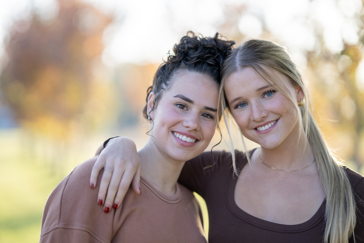 Best Friends Two teen girls huddle in closely together outside, with their arms around one another, as they pose for a portrait. They are both dressed casually and are smiling.