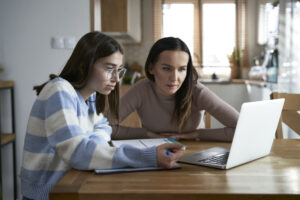 Mother with teenage daughter working on a laptop on a dining room table.
