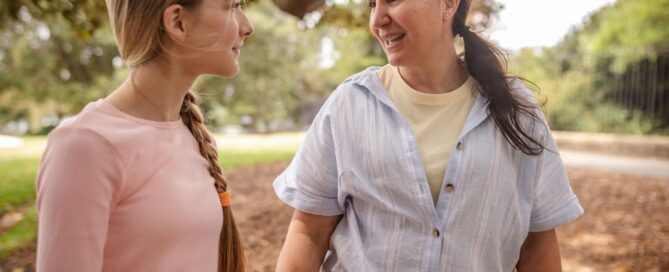 A smiling woman and a young teenage girl with a long braid look at each other and talk while walking outdoors in a park.