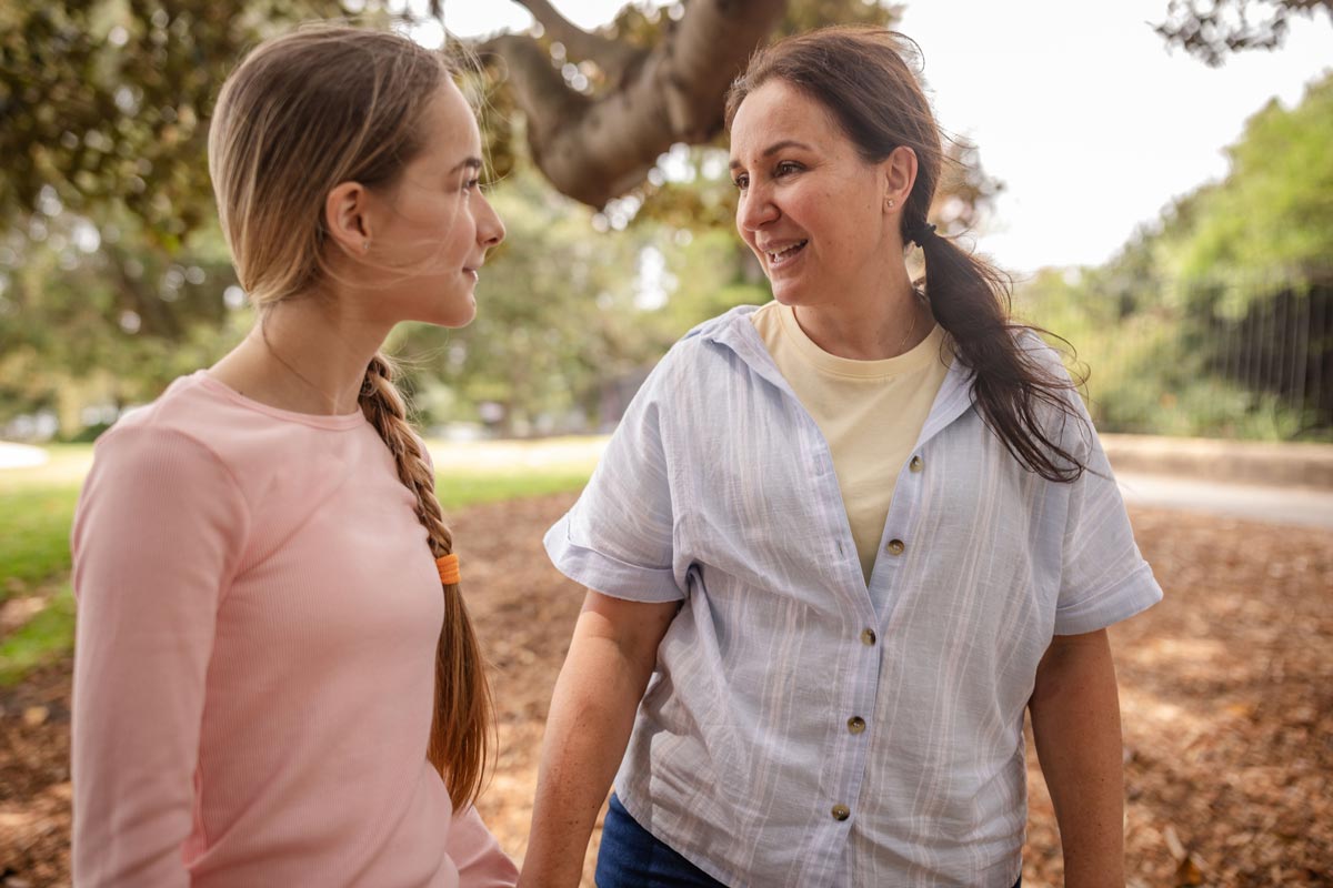 A smiling woman and a young teenage girl with a long braid look at each other and talk while walking outdoors in a park.