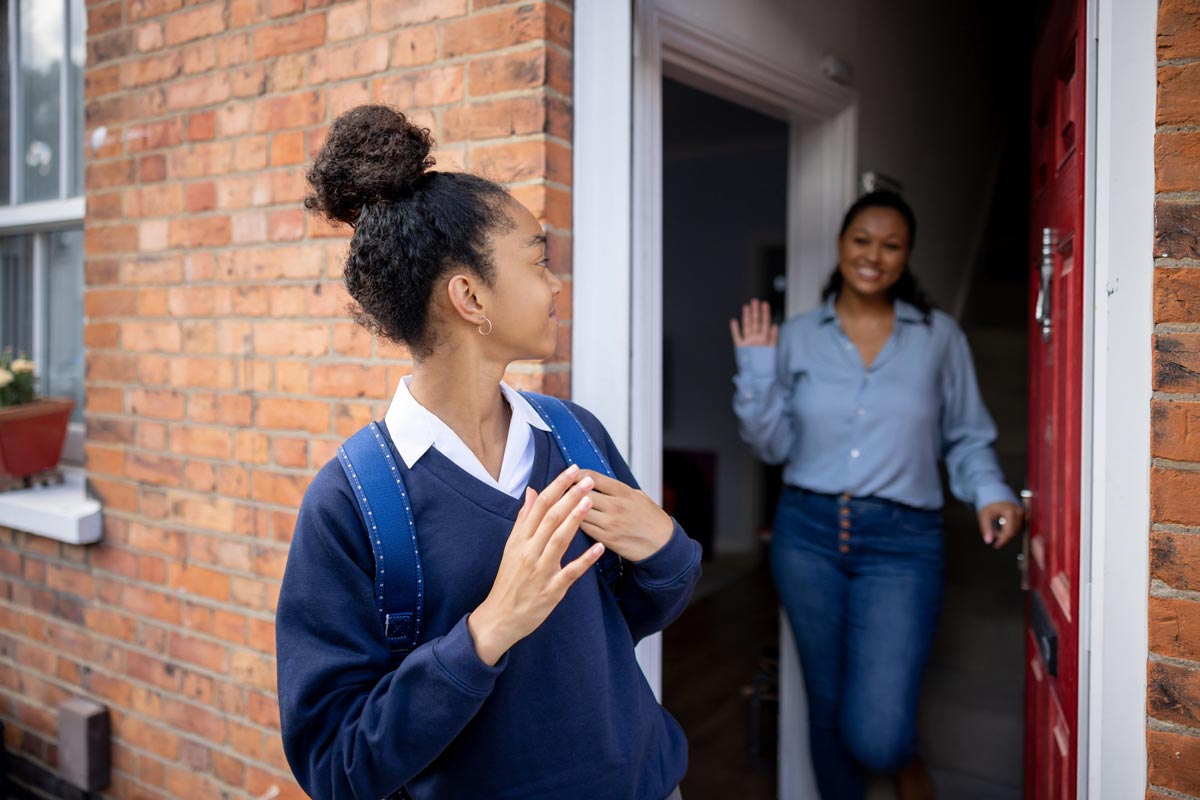 A teenage girl with a backpack waves goodbye as she walks away from a brick house, while a smiling woman waves back from the open doorway.