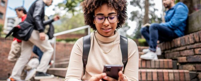 A young Black woman with curly hair and glasses sits on outdoor brick steps, smiling as she looks at her smartphone.
