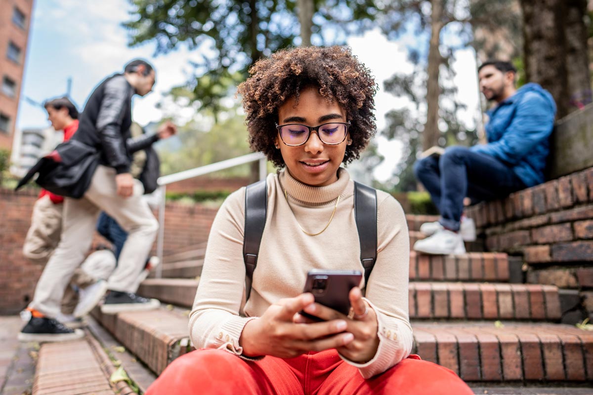 A young Black woman with curly hair and glasses sits on outdoor brick steps, smiling as she looks at her smartphone.