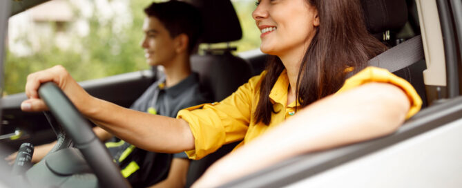 A smiling woman in a yellow shirt drives a car, with a young teenage boy sitting in the passenger seat beside her.