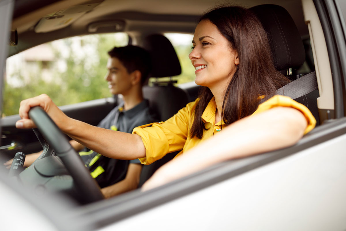 A smiling woman in a yellow shirt drives a car, with a young teenage boy sitting in the passenger seat beside her.