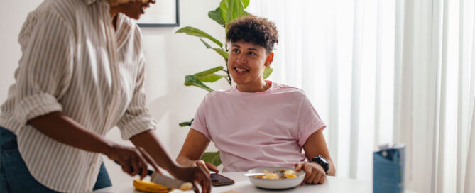 A woman with curly hair slices a banana at a dining table while talking to a teenage boy who is sitting and eating breakfast.