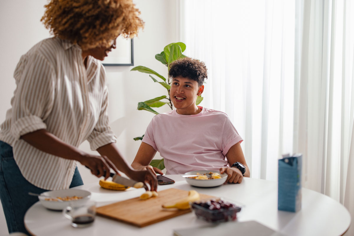 A woman with curly hair slices a banana at a dining table while talking to a teenage boy who is sitting and eating breakfast.
