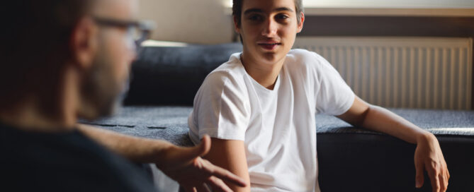 A teenage boy in a white t-shirt sits on the floor and listens to a man gesturing while talking in a living room.