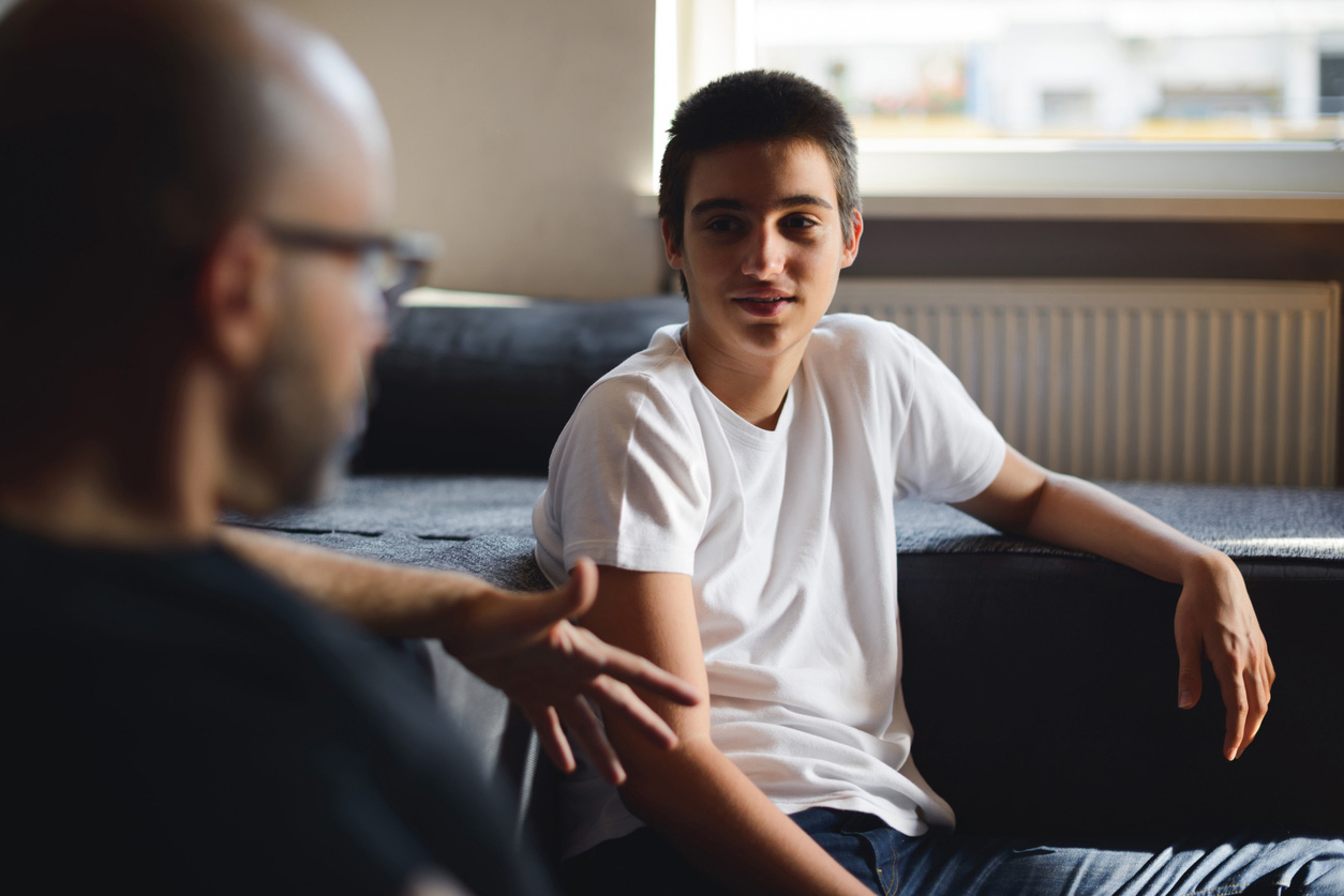 A teenage boy in a white t-shirt sits on the floor and listens to a man gesturing while talking in a living room.