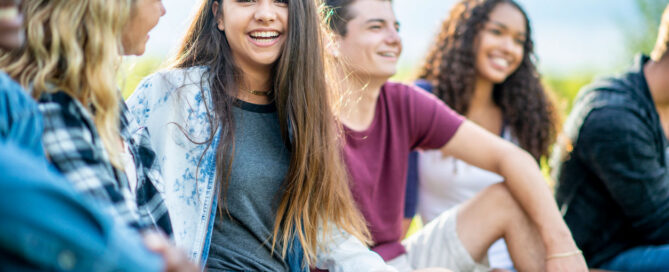 A group of diverse teenagers sit on the grass outdoors while a smiling girl in the center looks at the camera.