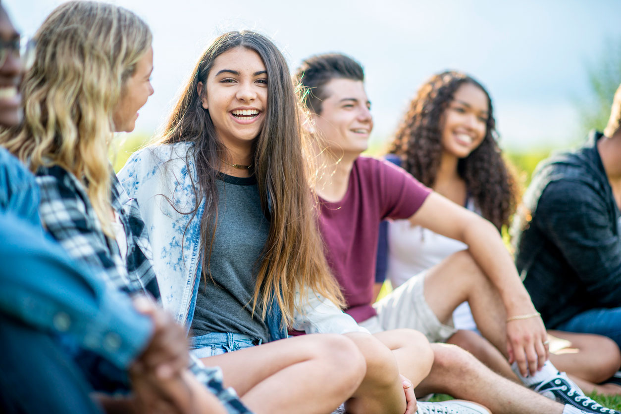 A group of diverse teenagers sit on the grass outdoors while a smiling girl in the center looks at the camera.