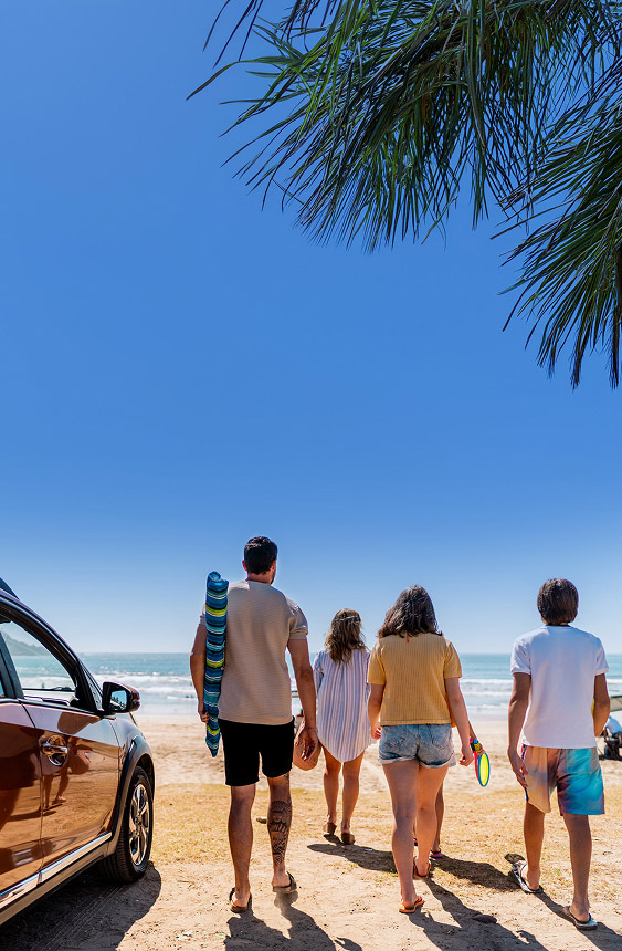 Rear view of four people walking from a parked car toward a sunny beach, framed by palm leaves at the top.