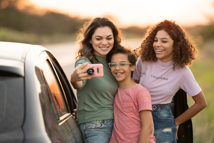 A woman, boy with glasses, and teenage girl smile while taking a smartphone selfie next to a car at sunset.